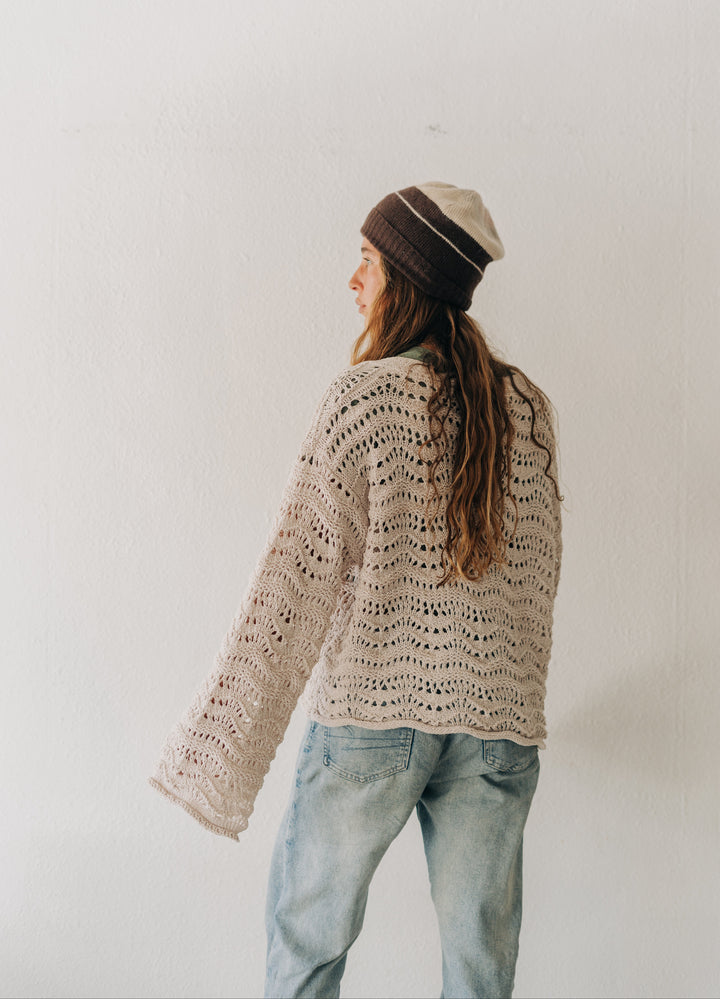 A woman wearing a cream-colored, open-knit crochet sweater, light-wash blue jeans, and a brown and beige striped beanie hat, standing against a plain white wall.