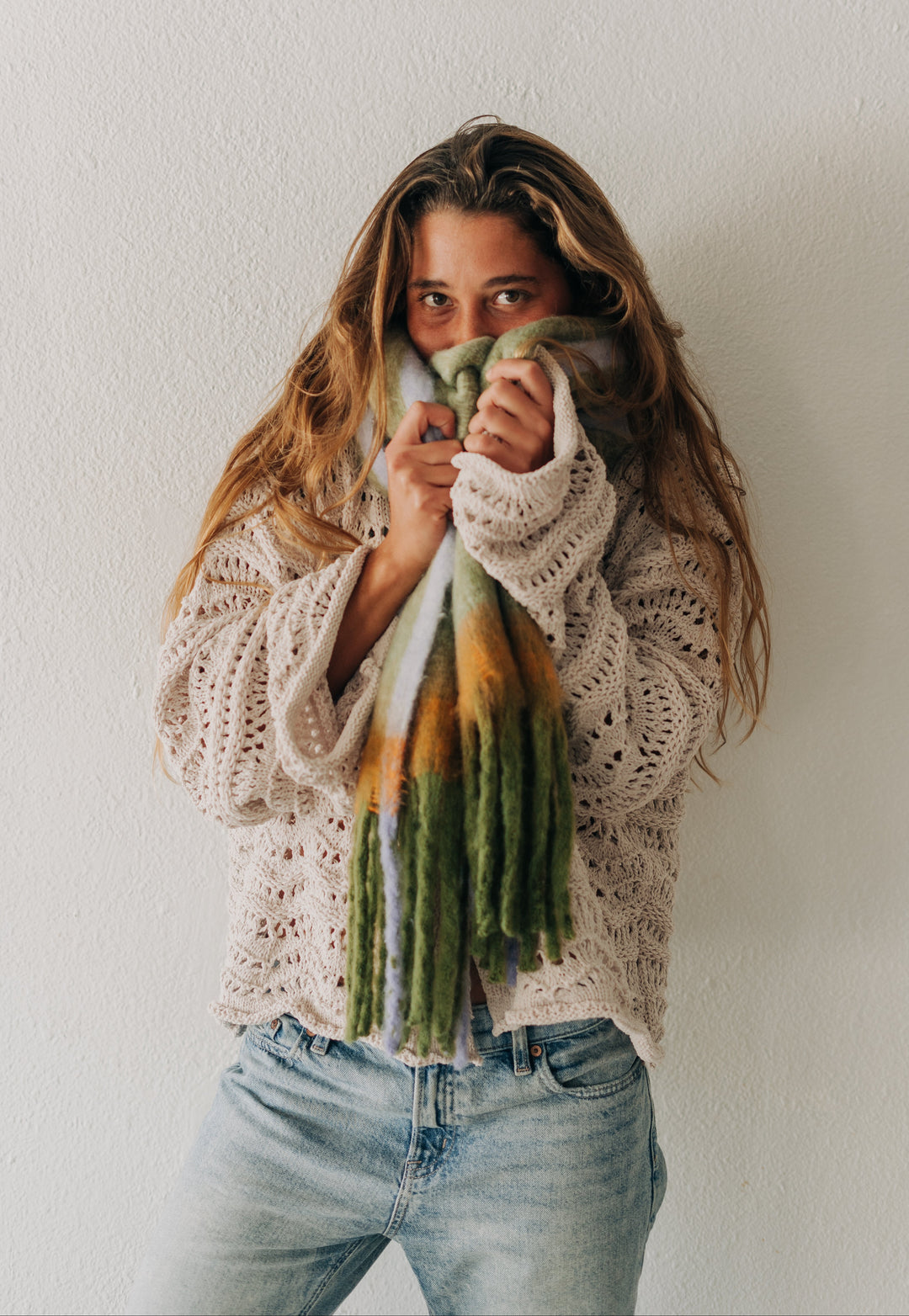 A woman wearing a cream-colored open-knit sweater and light-wash blue jeans, wrapped in a green, white, and orange tie-dye knit scarf, posing against a textured off-white wall.