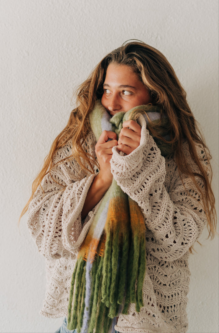 A woman with long light brown hair wearing a cream-colored crochet knit sweater and a green, yellow, and white gradient knit scarf, standing against a textured off-white wall.