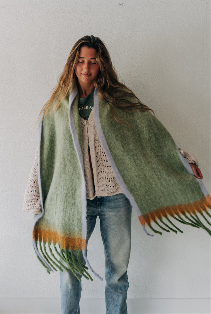 A woman wearing a cream knit sweater and blue jeans stands against a white wall, wrapped in a large sage green knit shawl with orange and green fringe details.
