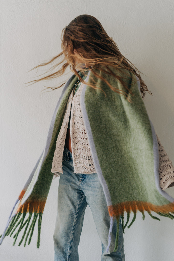 A woman wearing a light green knit shawl with orange and lavender fringe details, draped over her shoulders, paired with a cream crochet top and blue jeans against a plain white wall.