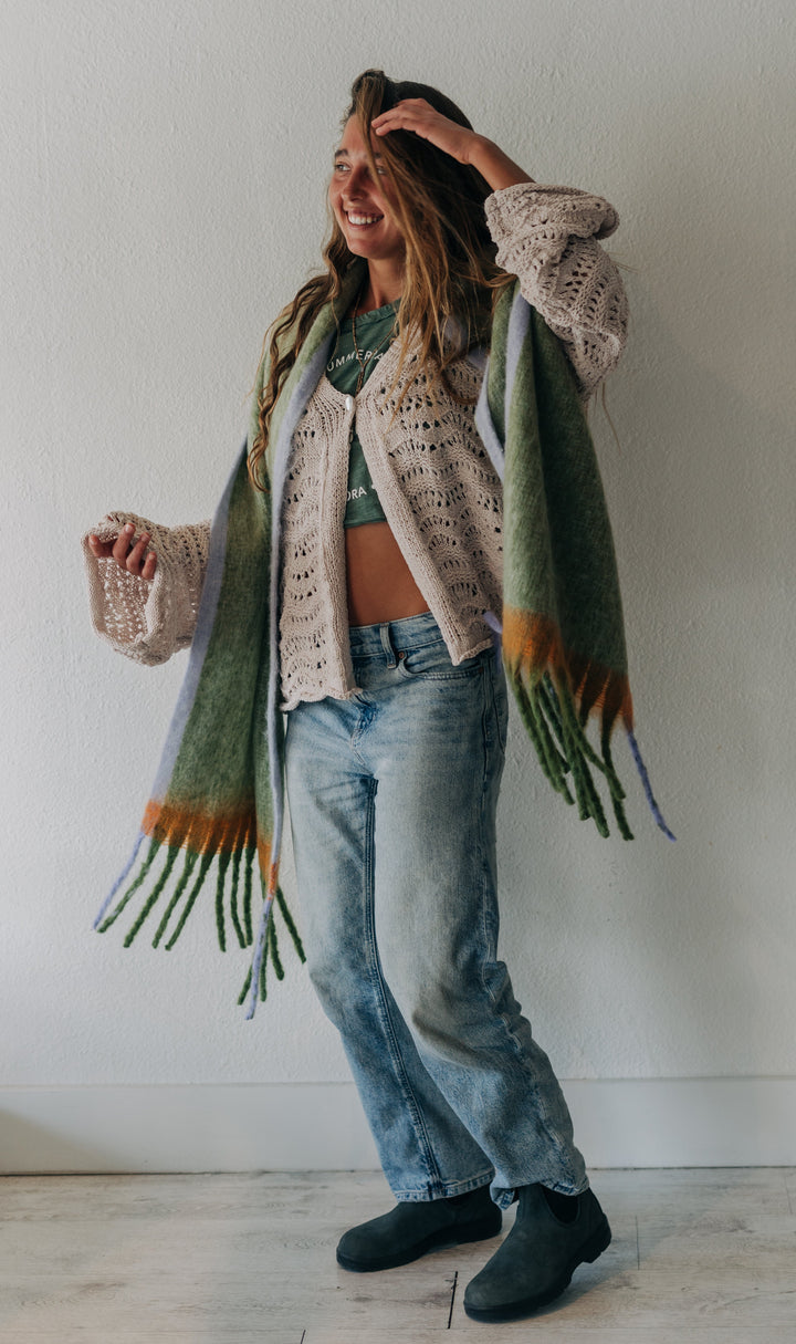 A woman stands against a plain off-white wall wearing a cream crochet cardigan, a green and orange fringed knit scarf, blue jeans, and black boots.