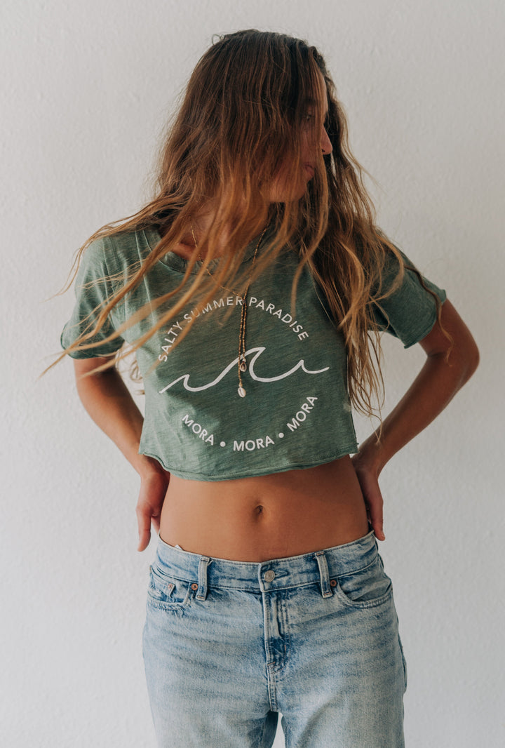 A woman with long light brown hair stands against a white wall wearing a cropped sage green graphic t-shirt and light wash blue jeans.