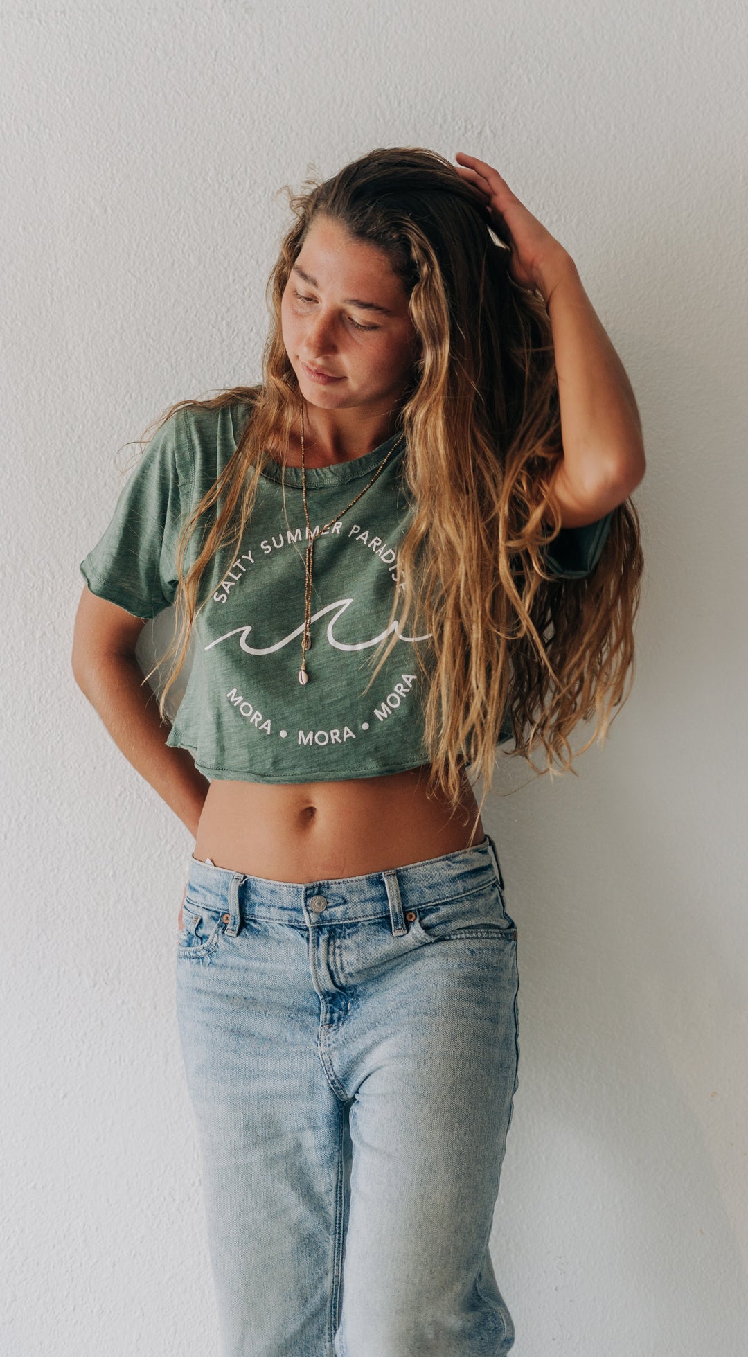 A young woman with long wavy light brown hair stands against a white wall wearing a sage green cropped graphic t-shirt and light wash blue jeans.