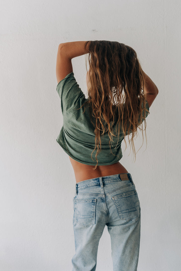 A person with long, wet, light brown hair stands with their back to the camera, wearing a green cropped t-shirt and light-wash blue jeans against a plain white wall.
