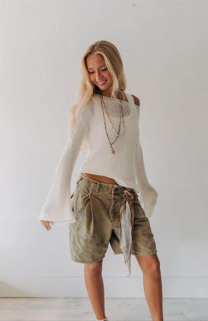 A woman modeling a white off-the-shoulder knit sweater and olive green denim shorts with a patterned fabric tie, standing against a plain white wall.