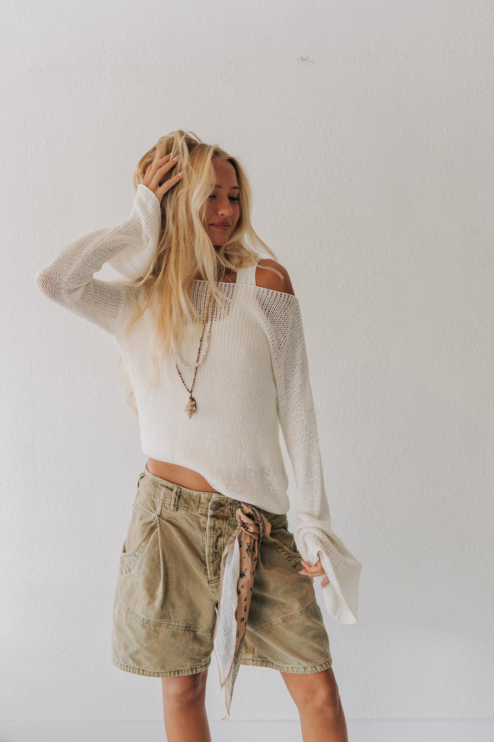A woman modeling a white off-the-shoulder knit sweater and an olive green utility mini skirt with a patterned tie belt, standing against a plain white wall.