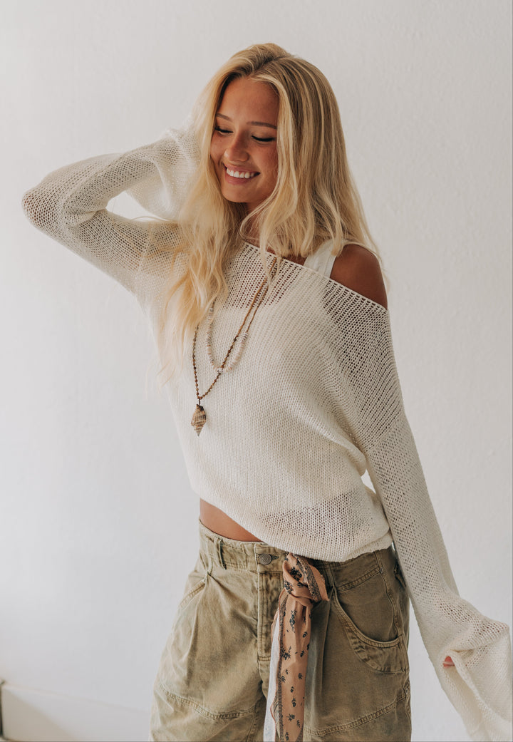 A woman with long blonde hair smiling while wearing a cream-colored, off-the-shoulder knit sweater and olive green cargo pants with a patterned scarf tied at the waist, set against a plain white wall.