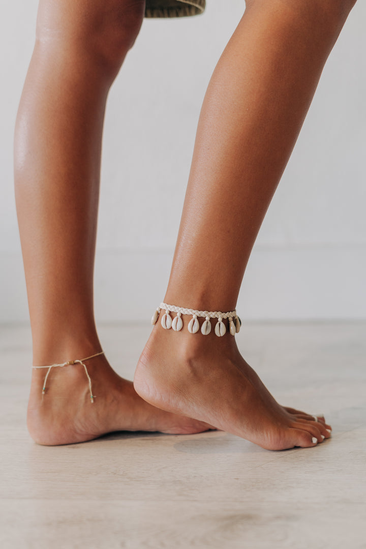A pair of delicate gold anklets, one with a simple chain and the other adorned with small white teardrop shell charms, shown on bare feet against a neutral light background.