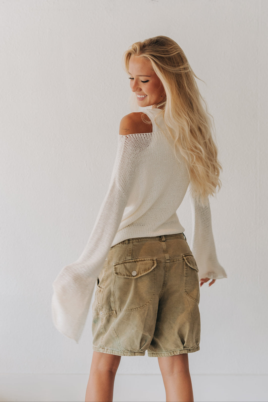 A woman with long blonde hair is shown from the back wearing a cream off-the-shoulder knit sweater and khaki cargo shorts against a plain white wall.