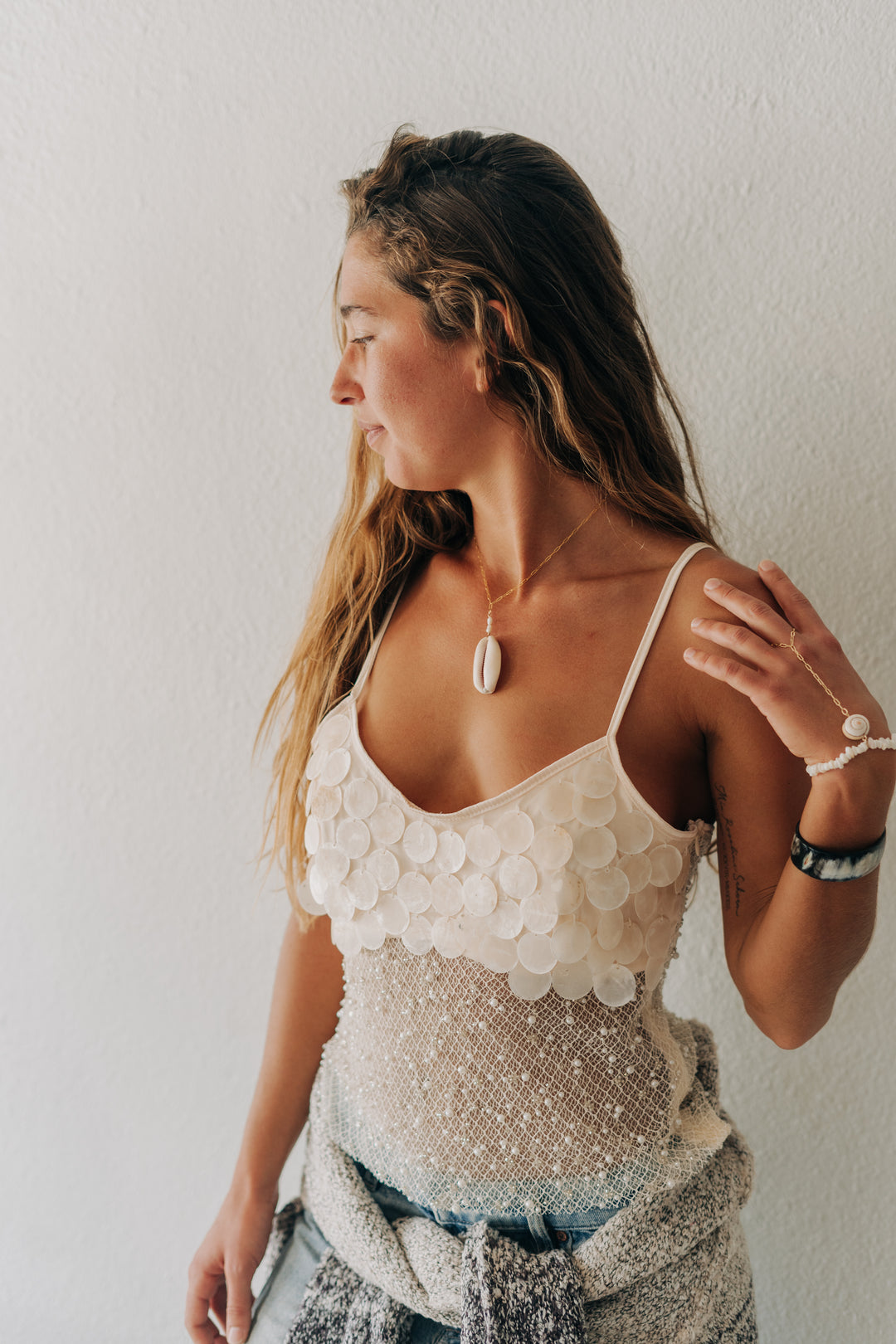 A woman wearing a bohemian-style cream sequin camisole top with scalloped edges and a shimmering sequined bodice, paired with a gray patterned skirt, standing against a textured off-white wall.