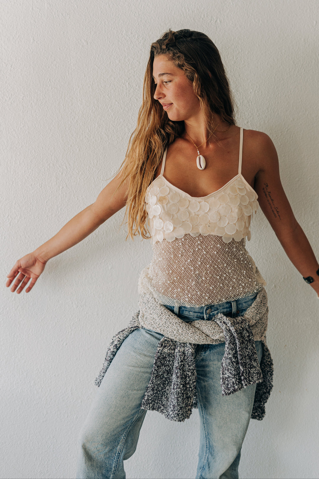 A woman posing against a white wall wearing a cream lace camisole top, light wash blue jeans, and a gray knit sweater tied around her waist.