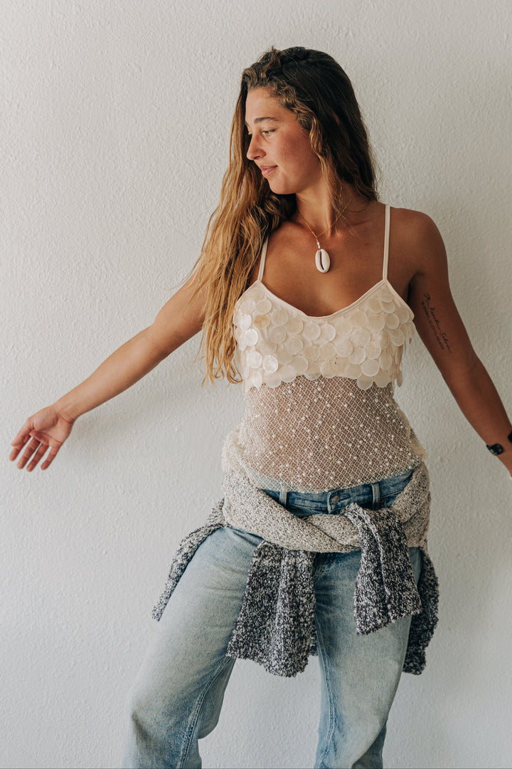 A woman posing against a white wall wearing a cream lace camisole top, light wash blue jeans, and a gray knit sweater tied around her waist.