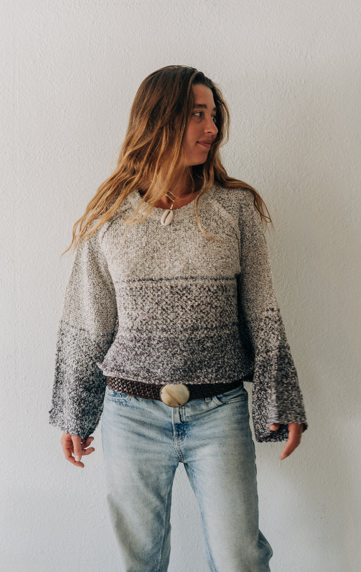A woman wearing a gray and white patterned knit sweater with bell sleeves, paired with light-wash blue jeans and a brown belt, standing against a plain white wall.