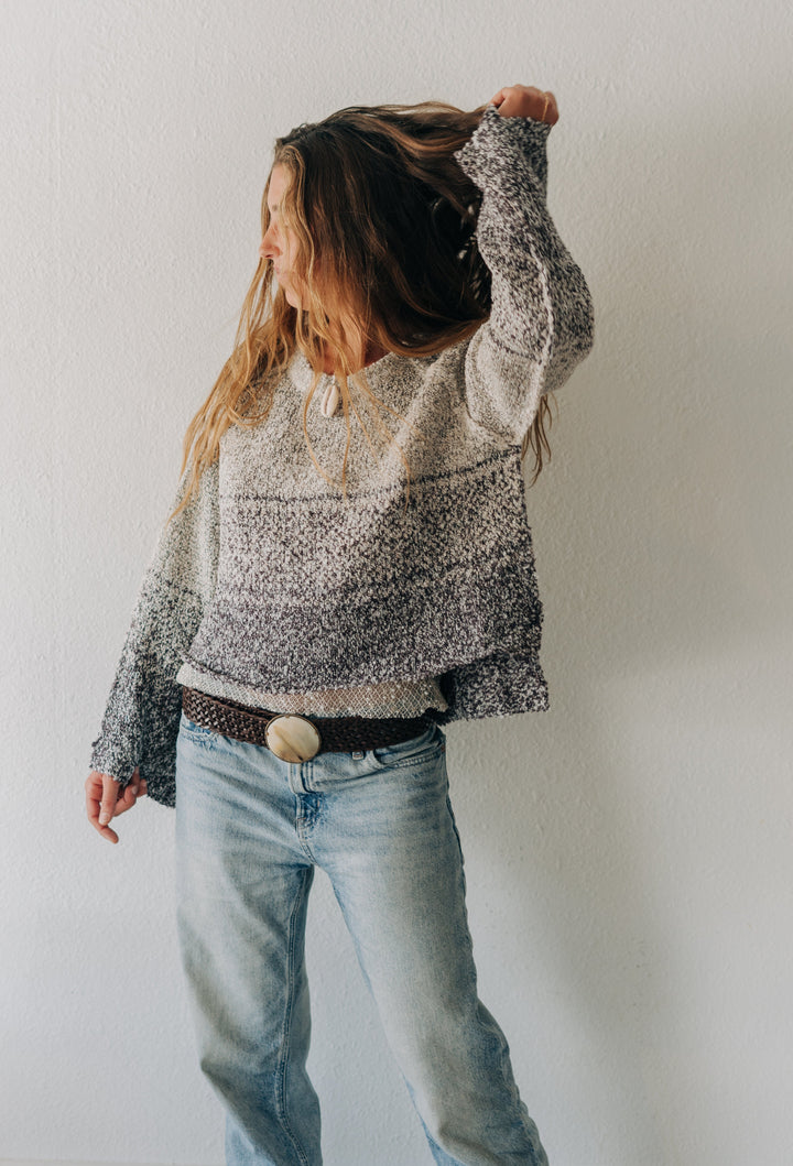 A woman wearing a gray and white marled knit sweater, light wash blue jeans, and a brown belt with a large round buckle, posing against a plain white wall.