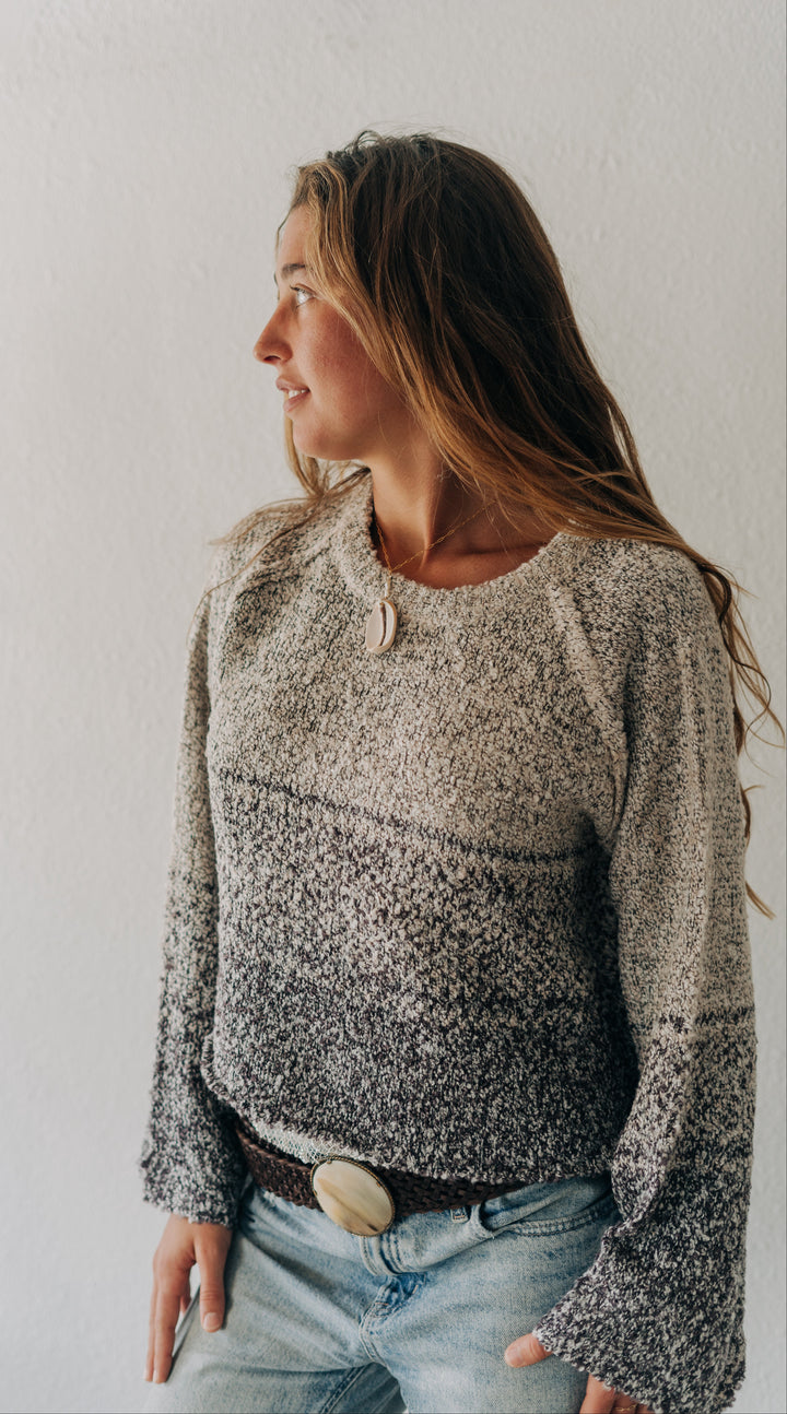 A woman wearing a long-sleeve knit sweater with a mottled gray and white pattern, paired with light blue jeans and a brown belt, standing against a plain white wall.