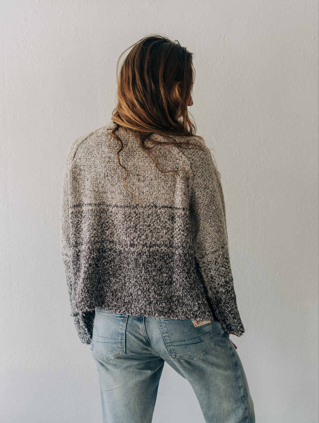 A woman viewed from behind wearing a textured, heathered knit sweater with a gradient from light beige to dark gray, paired with light-wash blue jeans against a plain white wall.