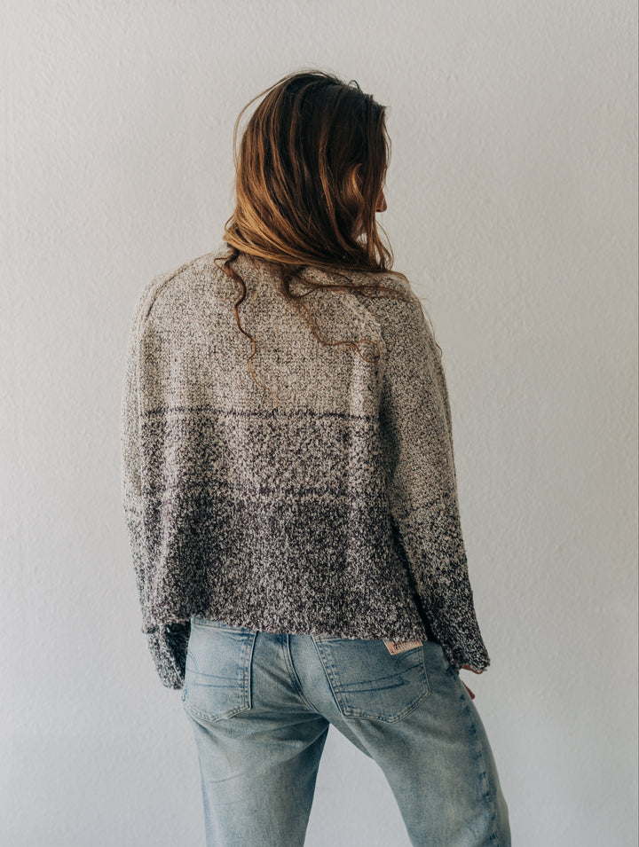 A woman viewed from behind wearing a textured, heathered knit sweater with a gradient from light beige to dark gray, paired with light-wash blue jeans against a plain white wall.