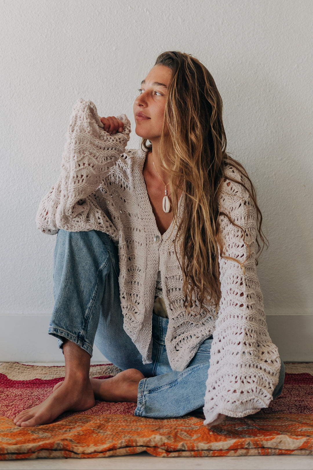 A woman sits cross-legged on a patterned rug against a white wall, wearing a cream open-knit cardigan, blue jeans, and barefoot.