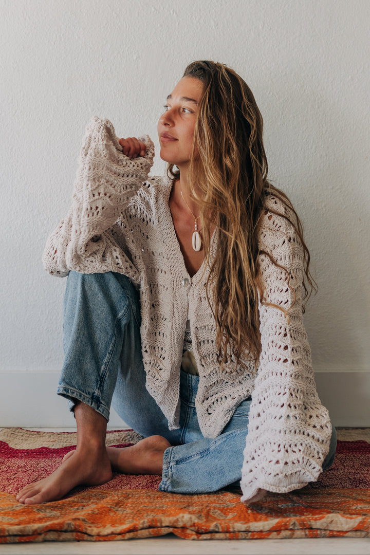 A woman sits cross-legged on a patterned rug against a white wall, wearing a cream open-knit cardigan, blue jeans, and barefoot.