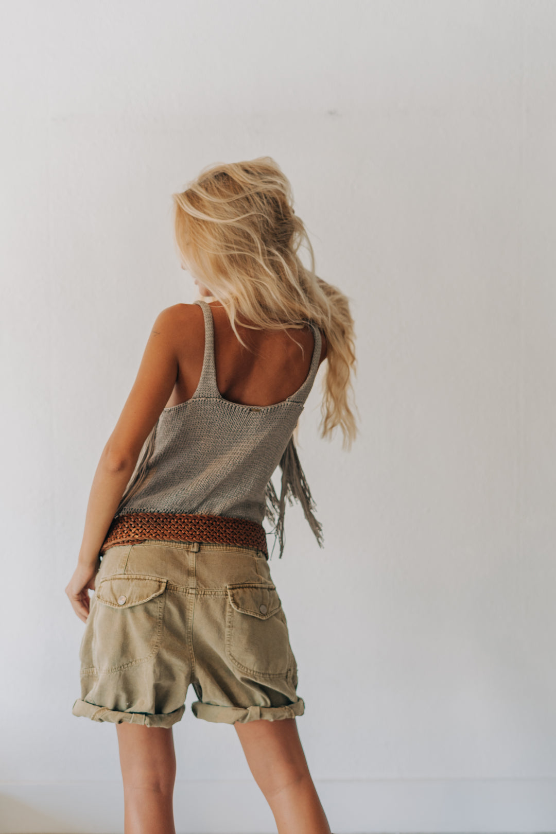 A young girl with long blonde hair is shown from behind, wearing a gray textured sleeveless top with fringe, khaki cargo shorts, and a brown patterned belt, standing against a plain white wall.