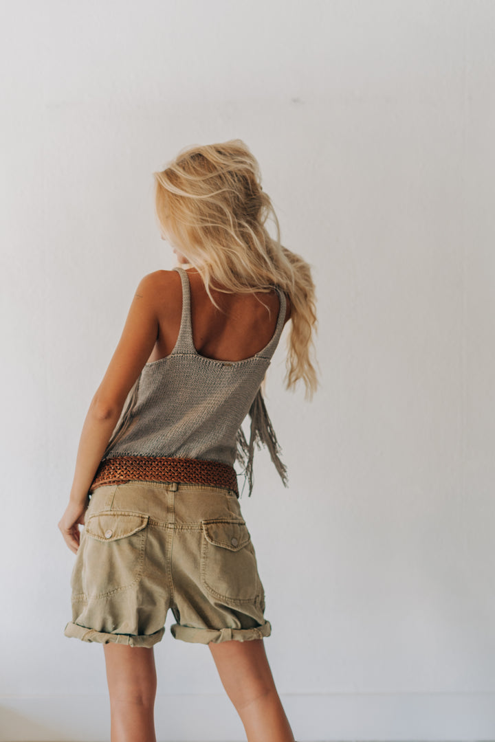 A young girl with long blonde hair is shown from behind, wearing a gray textured sleeveless top with fringe, khaki cargo shorts, and a brown patterned belt, standing against a plain white wall.