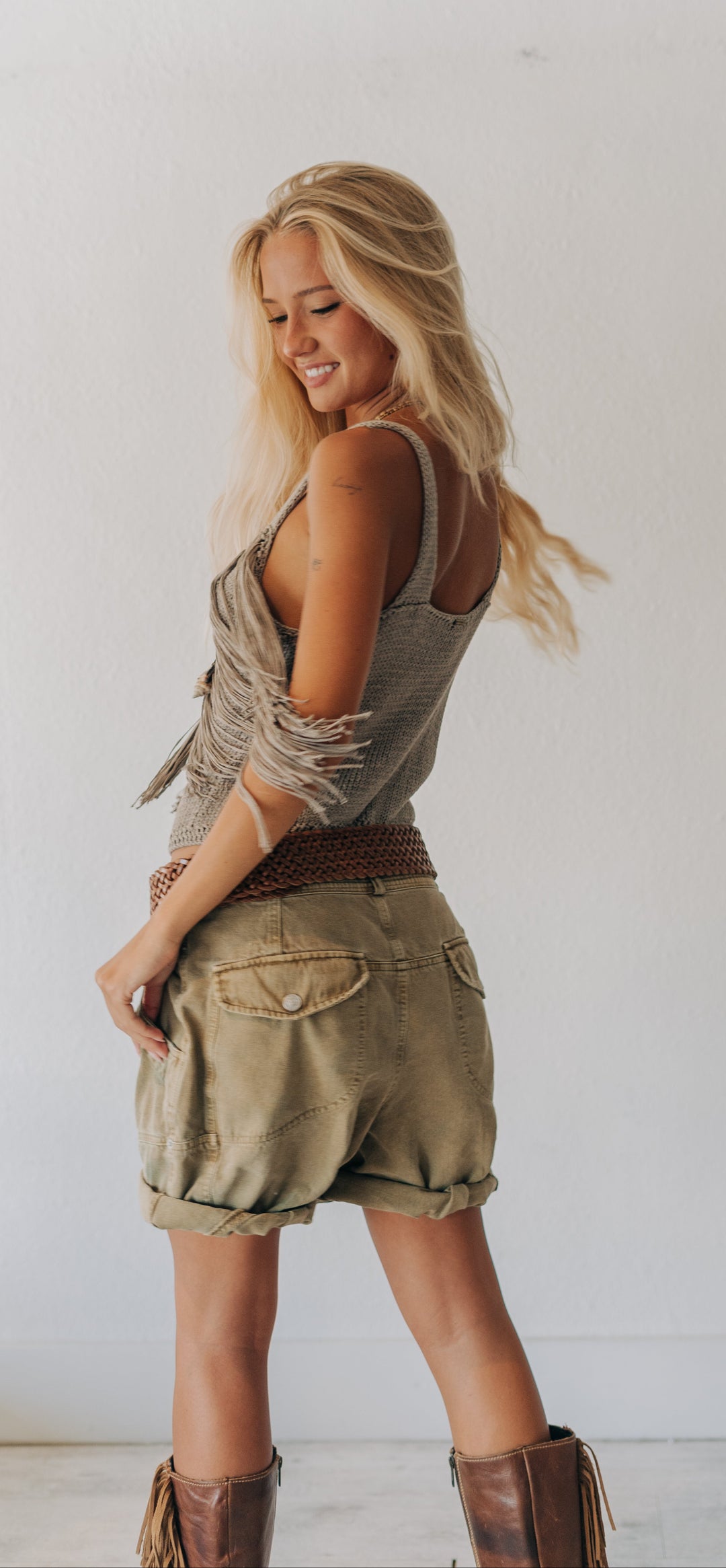 A blonde woman posing in a rustic, western-inspired outfit featuring an olive green fringed tank top, khaki cargo shorts, a brown belt, and brown leather boots, set against a plain white wall.