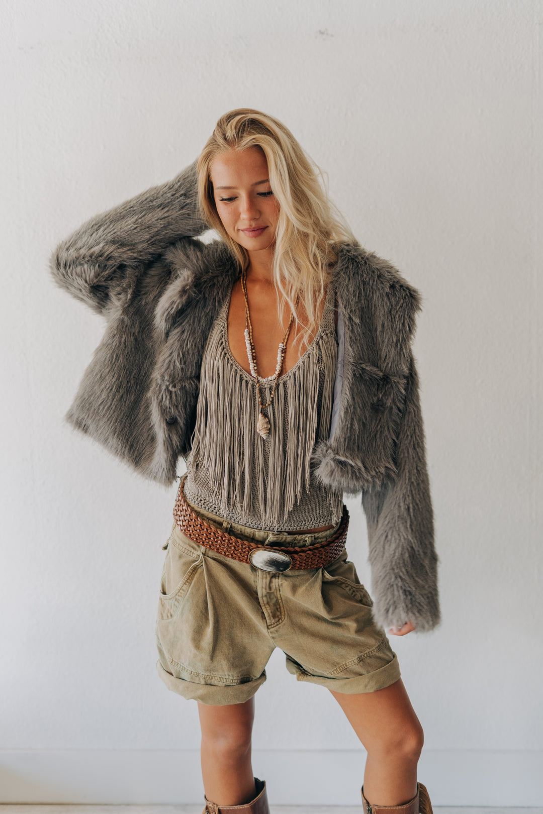 A woman modeling a bohemian-style outfit featuring a gray faux fur jacket, a beige fringe top, khaki shorts, and a brown woven belt, posed against a plain white wall.
