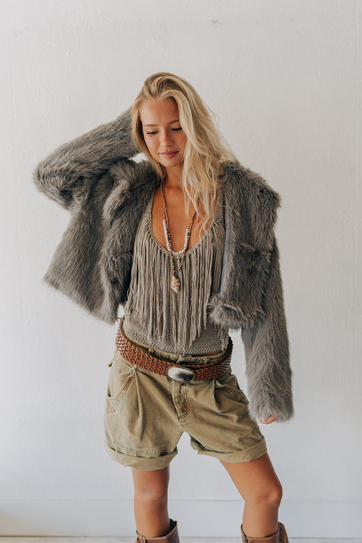 A woman modeling a bohemian-style outfit featuring a gray faux fur jacket, a beige fringe top, khaki shorts, and a brown woven belt, posed against a plain white wall.