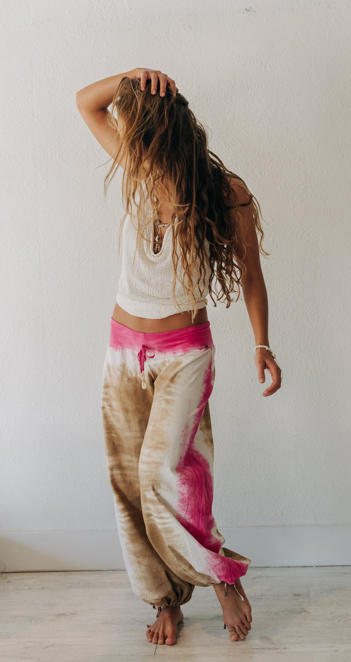 A woman with long wavy brown hair poses in a white crop top and tie-dye sweatpants featuring beige, white, and vibrant pink. She is standing barefoot in a minimalist room with a white wall and light wood floor.