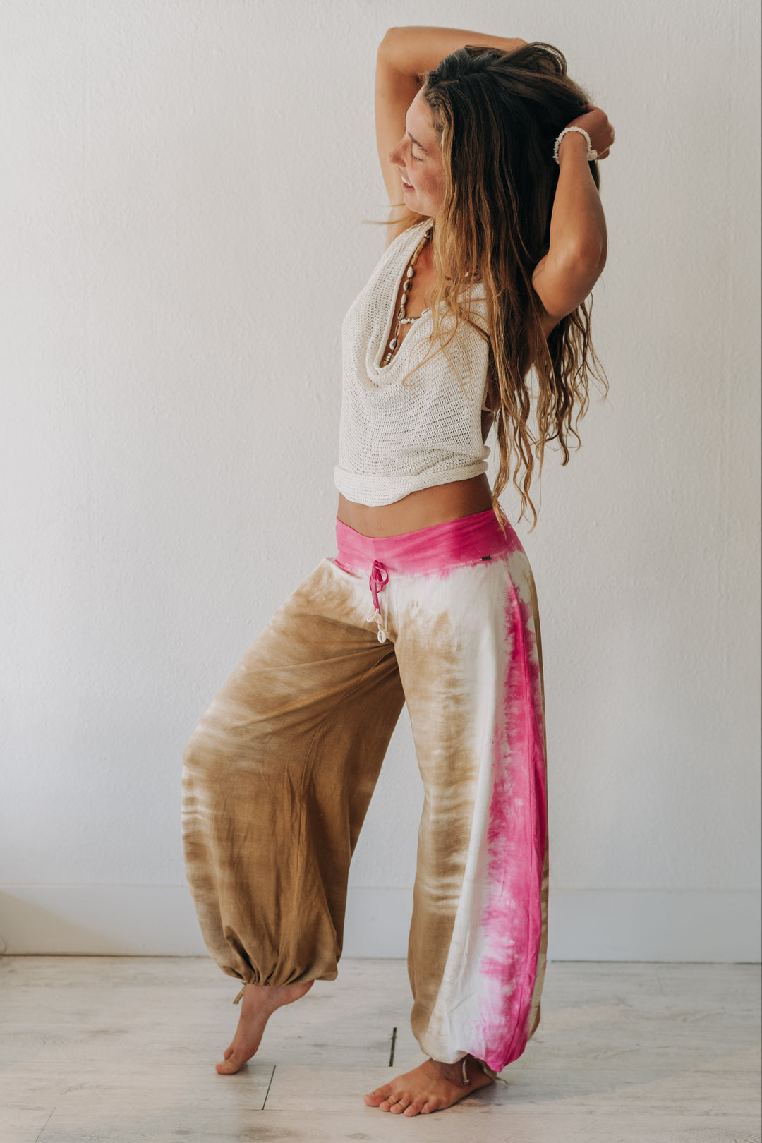 A woman posing in a room wearing tie-dye harem pants with a brown, white, and pink pattern, paired with a white knit crop top.