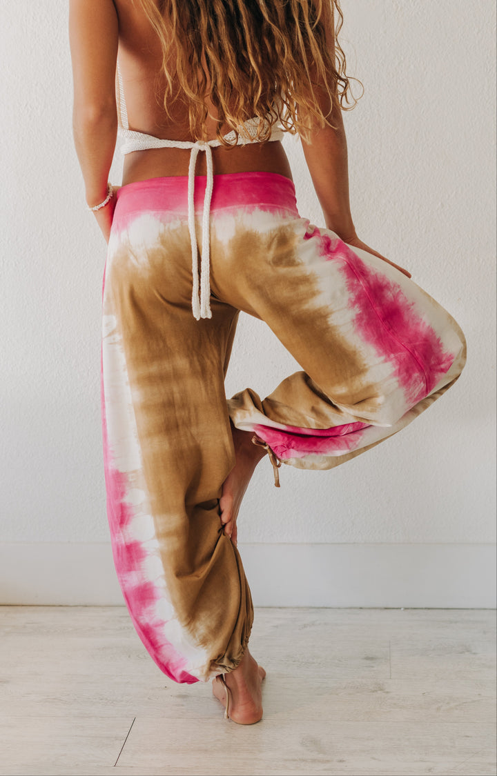 A woman wearing loose, tie-dye harem-style pants in brown, pink, and white, shown from the back against a plain white wall and light wood floor.