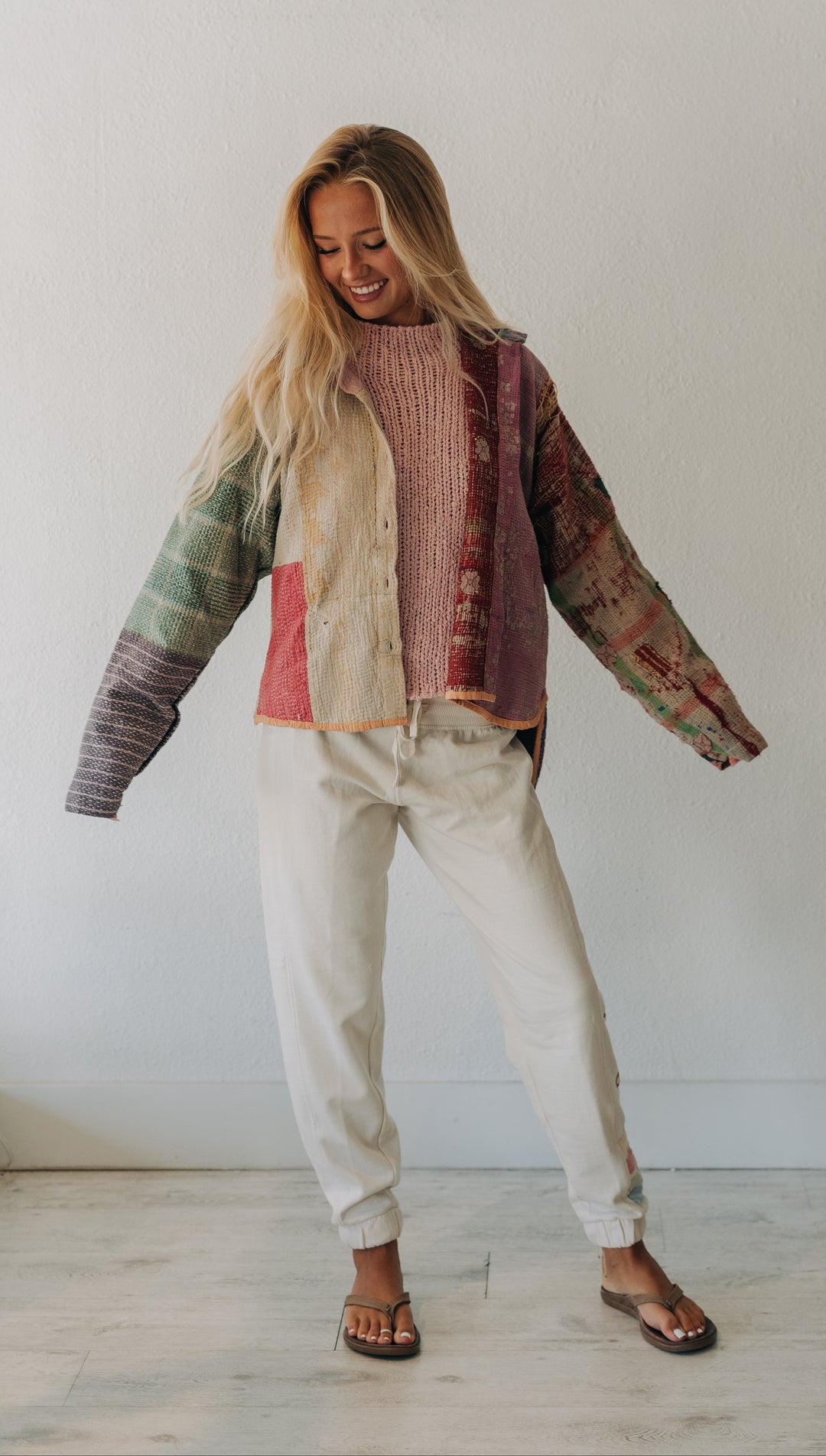 A woman modeling a colorful patchwork knit cardigan with cream, pink, green, and brown tones, paired with off-white pants and dark sandals, standing against a plain white wall.