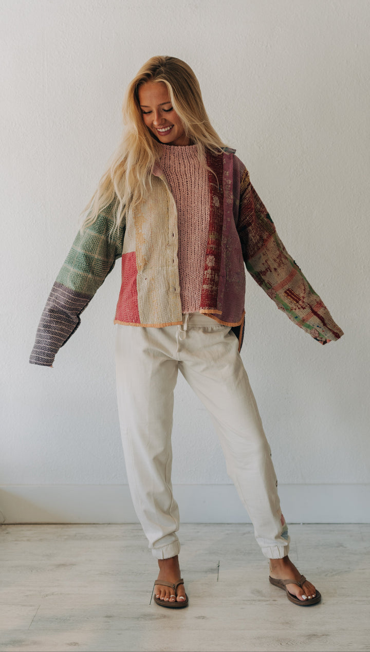 A woman modeling a colorful patchwork knit cardigan with cream, pink, green, and brown tones, paired with off-white pants and dark sandals, standing against a plain white wall.