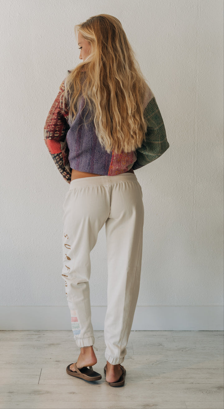 A woman with long blonde hair is shown from behind wearing cream-colored distressed jogger pants and a multi-colored patchwork knit sweater, standing against a plain white wall.
