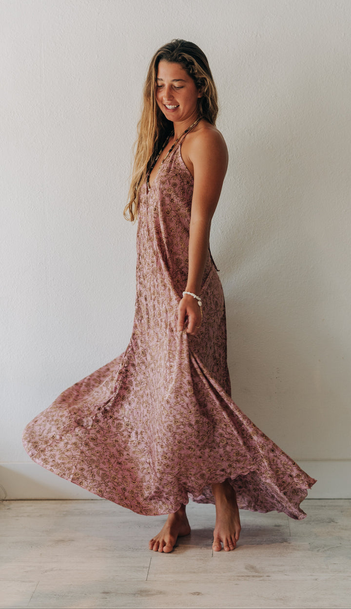A woman wearing a dusty rose floral print maxi dress, captured mid-spin as the dress flows around her bare feet against a plain white wall.