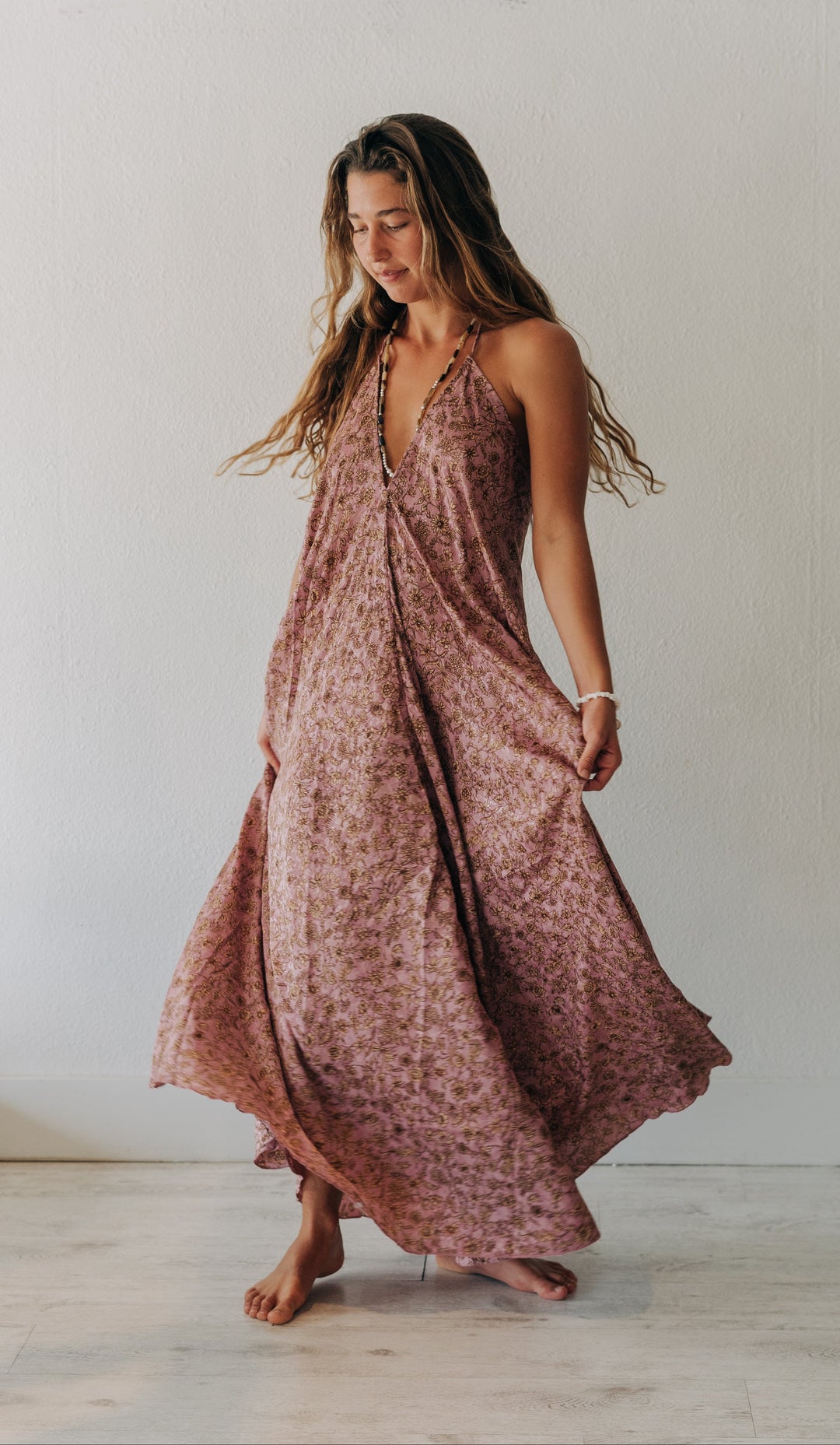 A woman wearing a flowing sleeveless maxi dress with a dusty rose and brown abstract print, standing barefoot in a bright, minimalist room with a white wall and light floor.