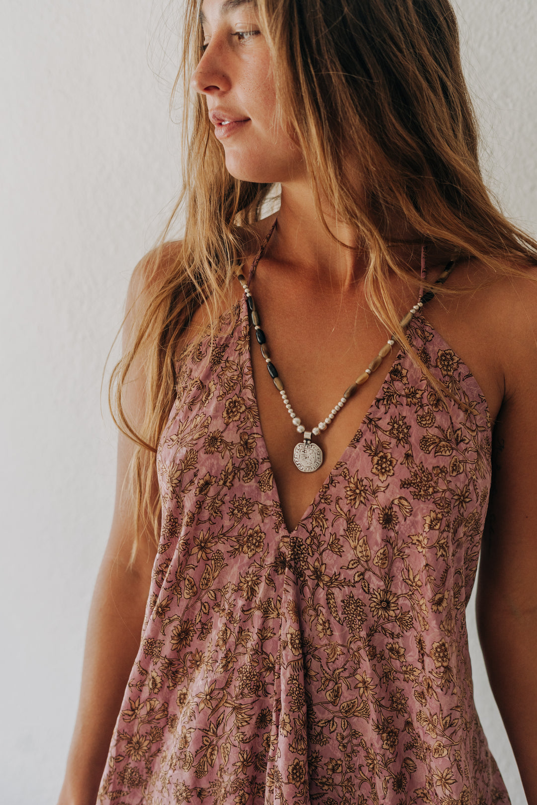 A woman wearing a dusty rose halter top with a brown floral pattern and a silver pendant necklace, shown in a close-up profile view against a white wall.