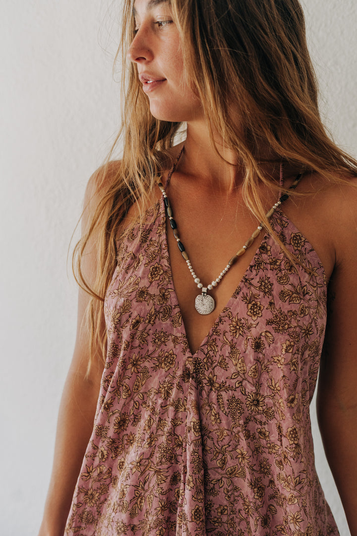 A woman wearing a dusty rose halter top with a brown floral pattern and a silver pendant necklace, shown in a close-up profile view against a white wall.