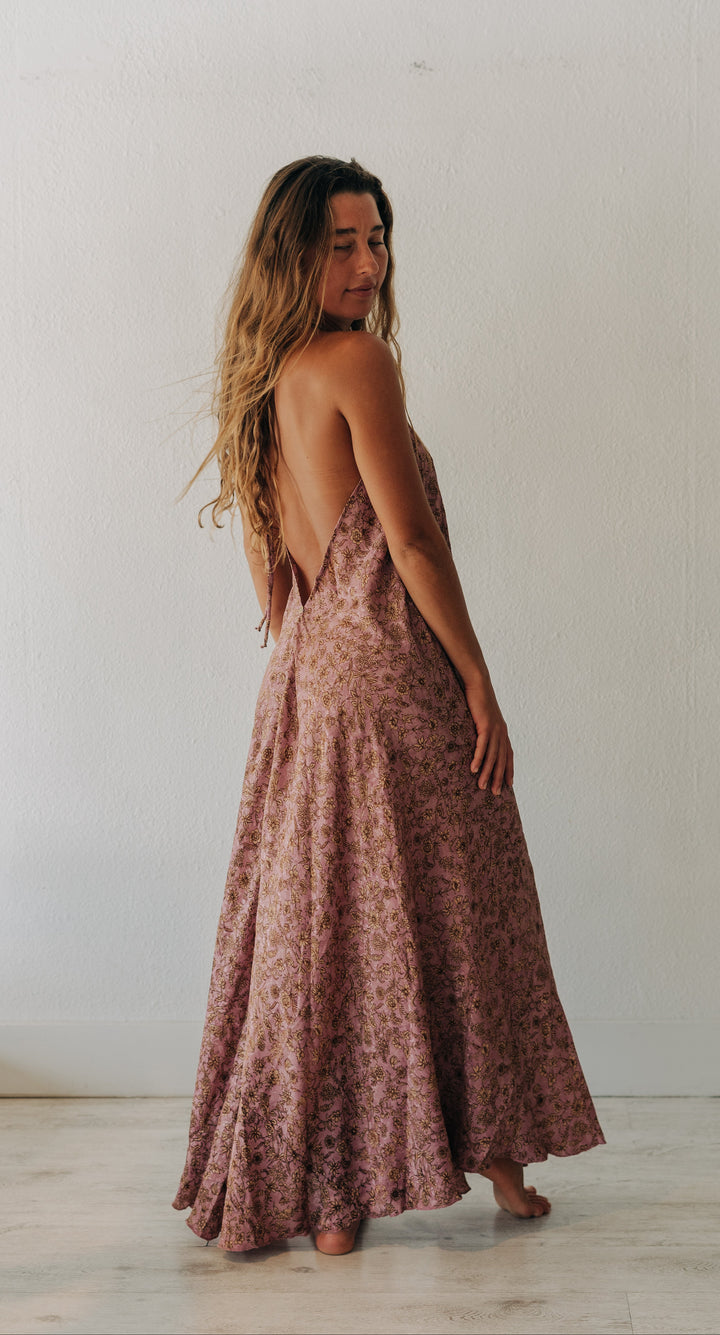 A woman wearing a sleeveless, backless maxi dress with a dusty rose and brown floral pattern, standing barefoot against a white wall.