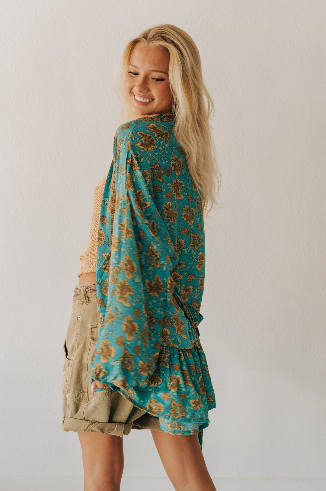 A woman with blonde hair is shown from the back wearing a teal and brown floral patterned shawl draped over her shoulders, paired with khaki cargo shorts, against a plain white wall.