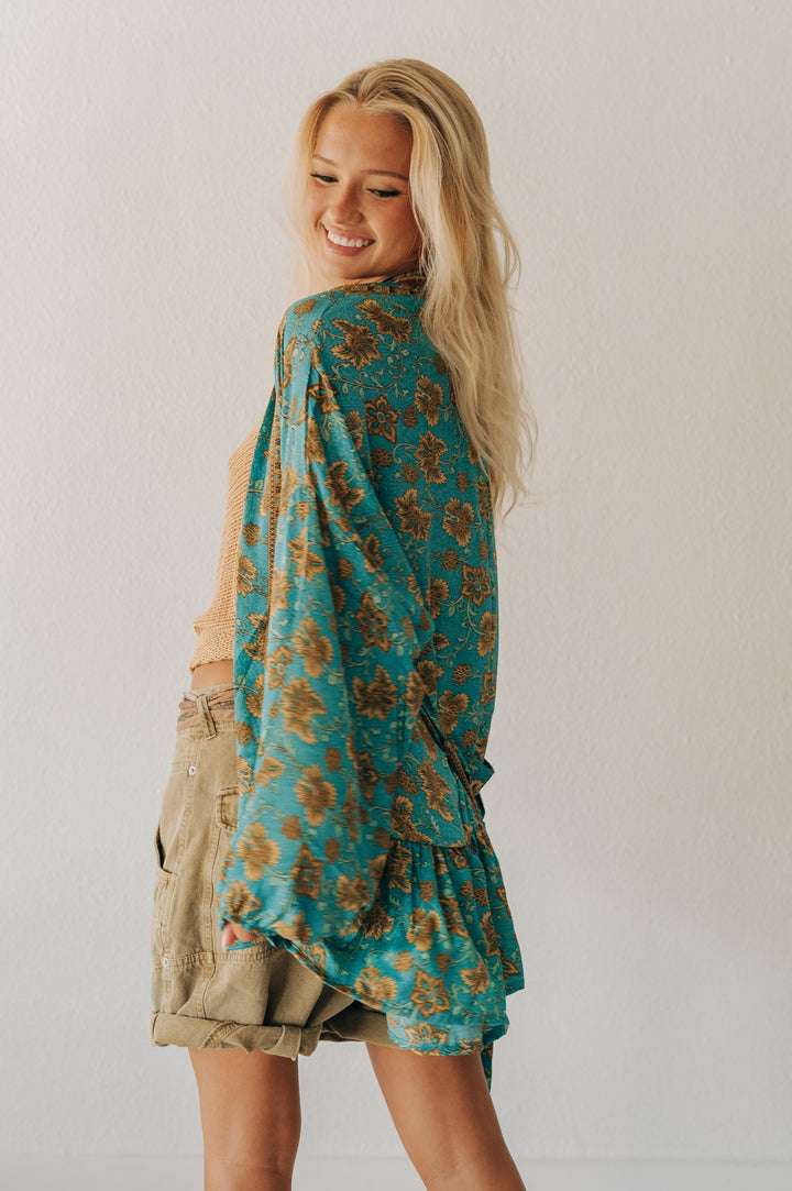 A woman with blonde hair is shown from the back wearing a teal and brown floral patterned shawl draped over her shoulders, paired with khaki cargo shorts, against a plain white wall.