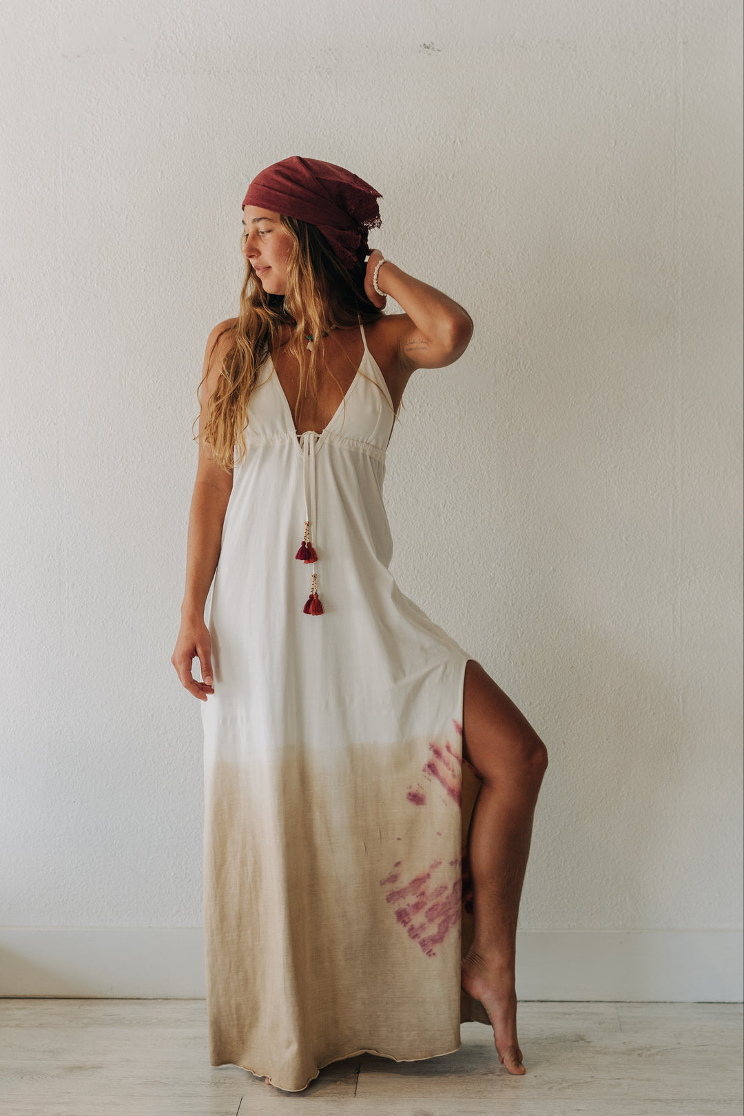 A woman wearing a white maxi dress with a beige gradient hem and red tassel ties, paired with a burgundy headwrap, standing in a minimalist room with a white wall and light wood floor.