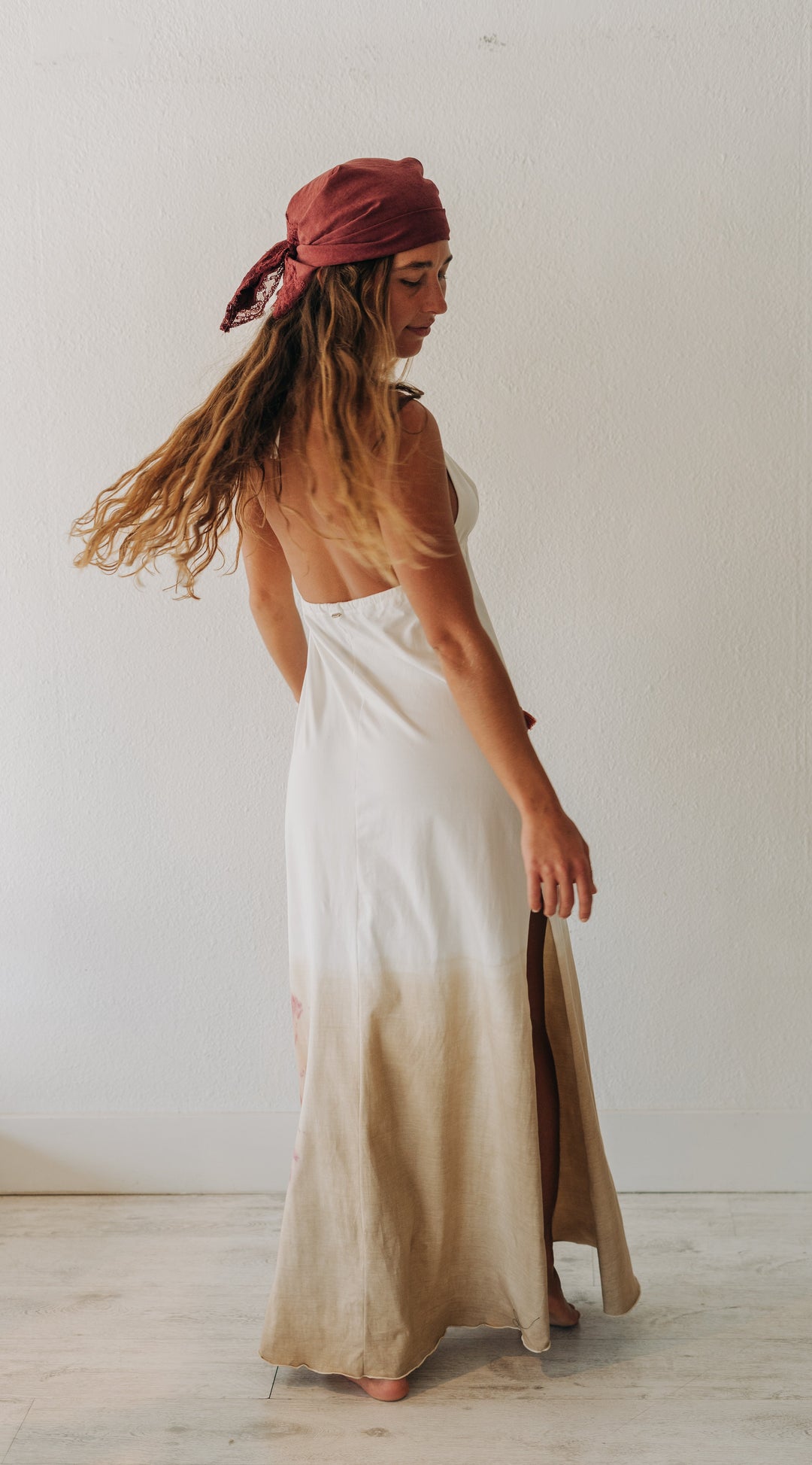 A woman wearing a sleeveless white maxi dress with a beige gradient hem and a rust red headscarf, standing in a minimalist white room.