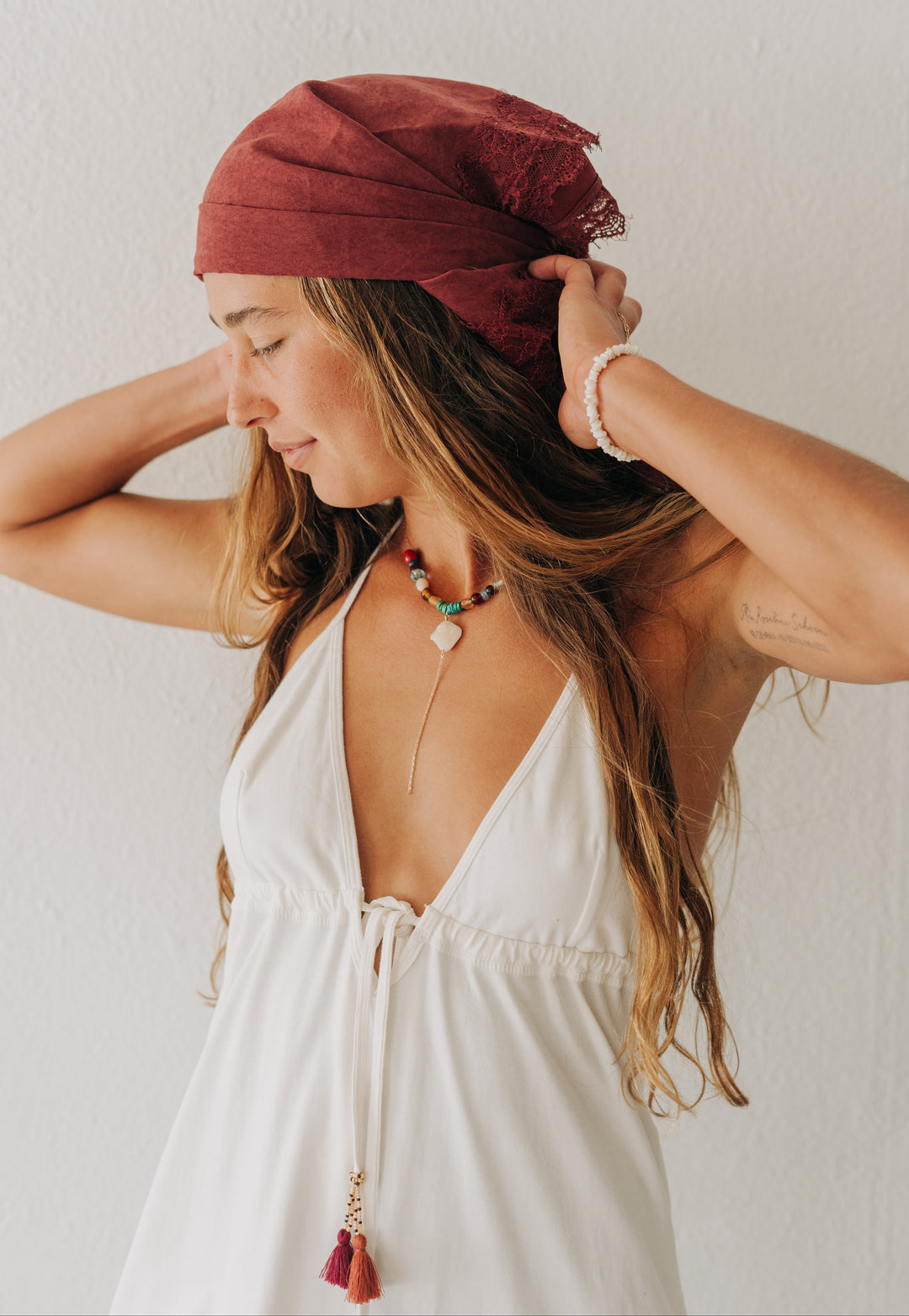 A woman wearing a rust-red linen headwrap and a white halter dress, adjusting the wrap against a plain white wall.