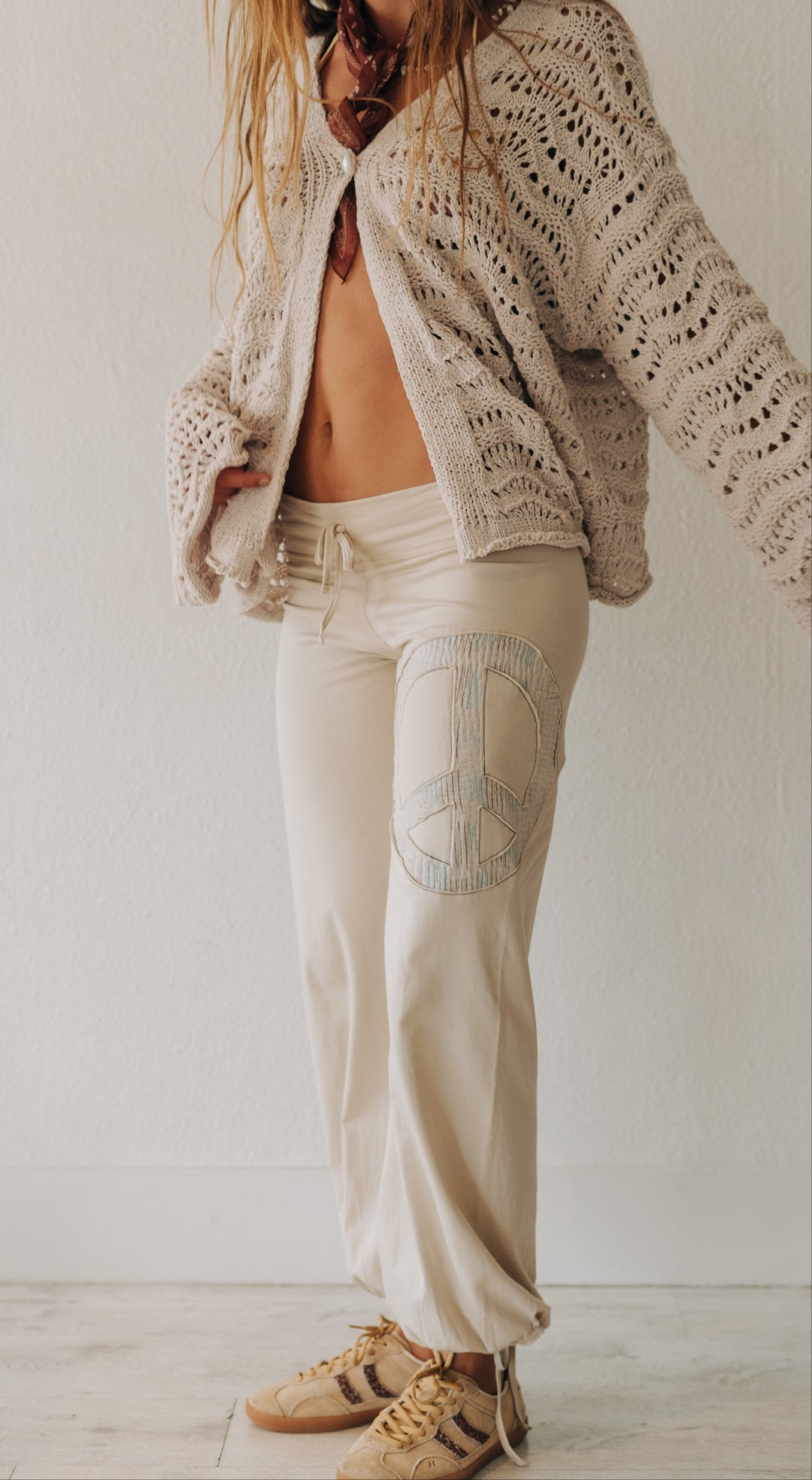 A woman wearing a cream-colored open-knit cardigan, matching cream drawstring pants with a peace sign patch on the thigh, and beige sneakers, standing against a plain off-white wall.