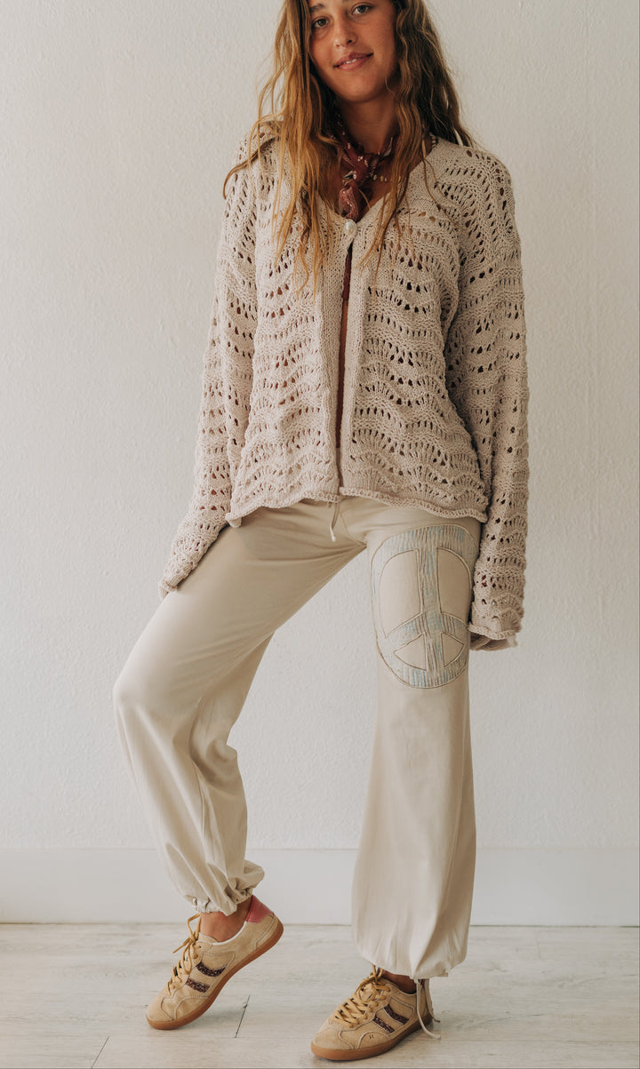 A woman modeling a cozy beige open-knit cardigan, cream wide-leg pants with a peace sign patch on the thigh, and tan sneakers, standing against a plain white wall.