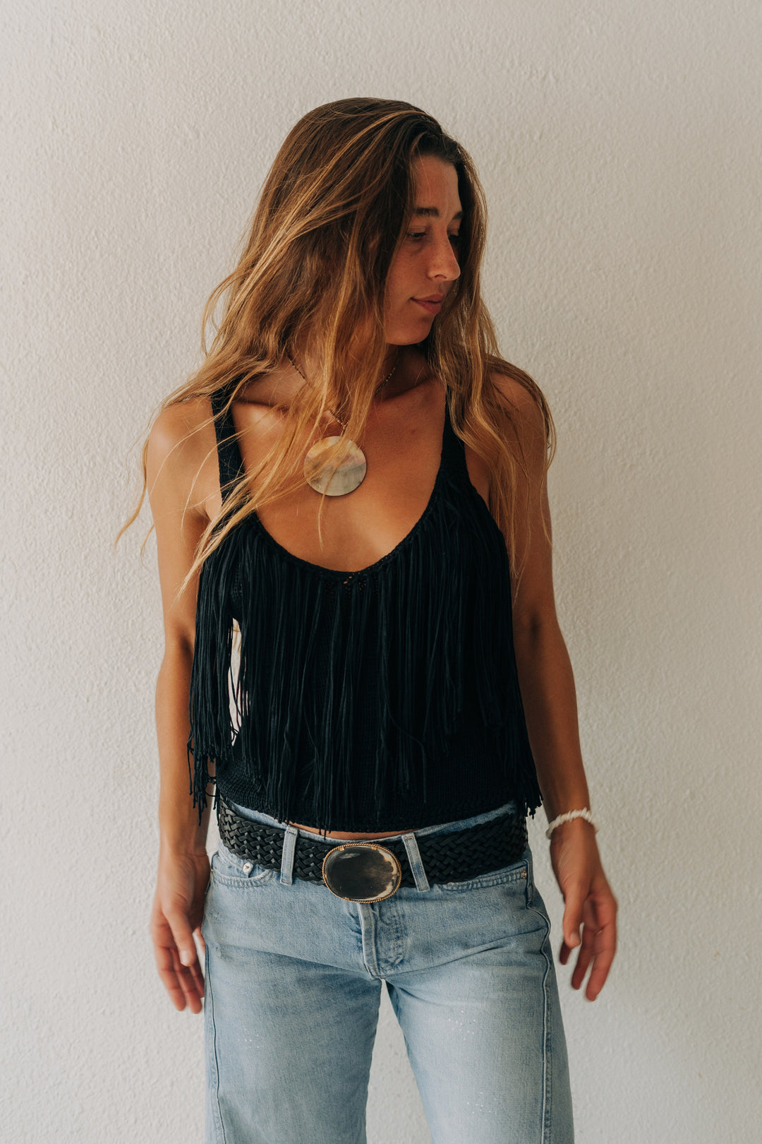 A woman with long light brown hair stands against a textured off-white wall, wearing a black fringed tank top, light-wash blue jeans, a black studded belt with a large silver buckle, and a chunky gold pendant necklace.