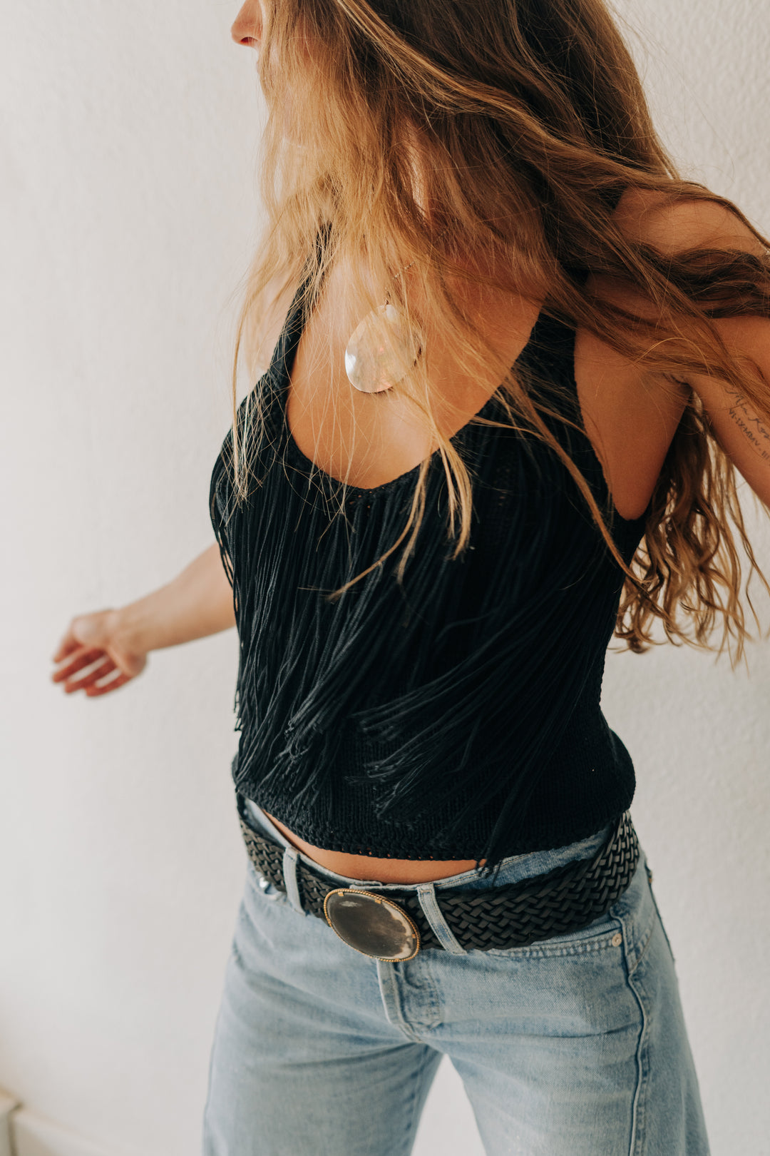 A woman wearing a black pleated tank top and light blue jeans, shown from a side-back angle against a plain white wall.