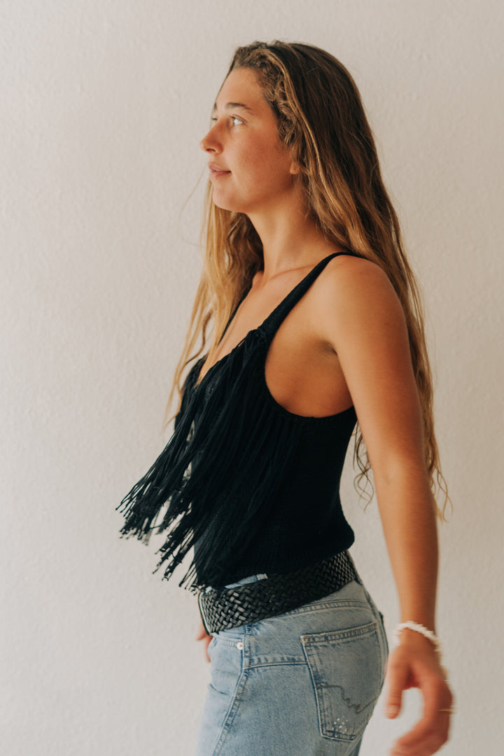 A woman in profile wearing a black fringed tank top and light blue jeans, standing against a plain off-white wall.